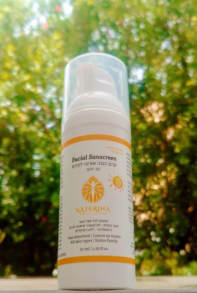 בקבוק של קרם הגנה לפנים SPF30 של קטרינה, עם עיצוב לבן ונחושת על גבעול צמחייה ירוקה ברקע.
