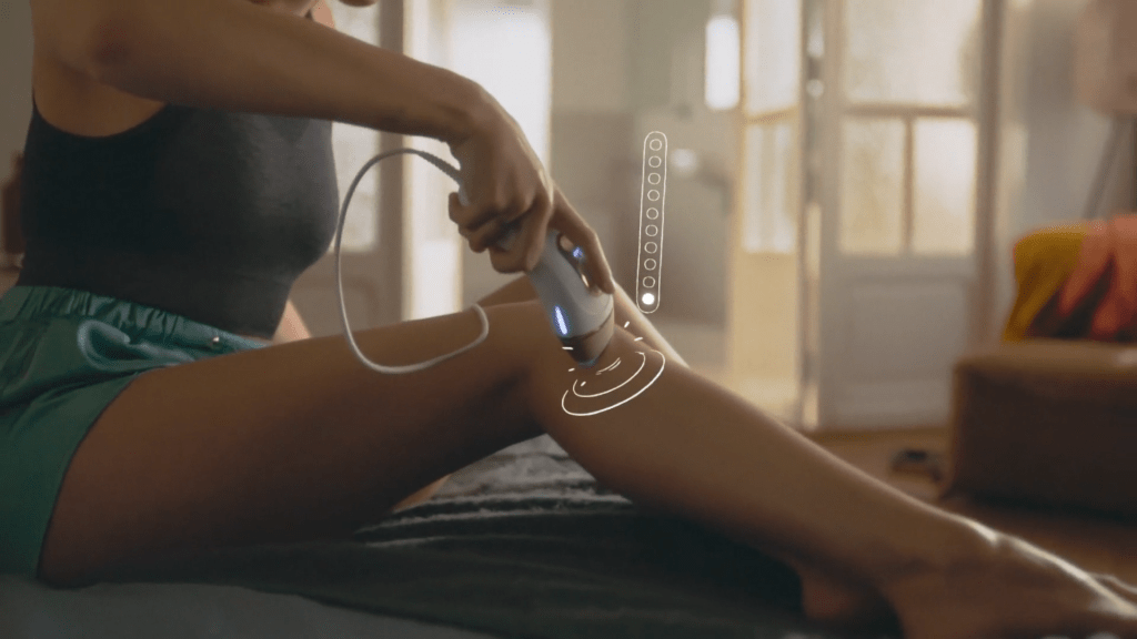 אישה משתמשת במכשיר הסרת שיער IPL על רגליה, יושבת בסלון עם ריהוט מודרני.