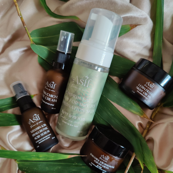 מגוון מוצרי קוסמטיקה טבעית של אסיף קוסמטיקה, כולל סרומים וקרמים, המונחים על בד רך עם עלים ירוקים ברקע.