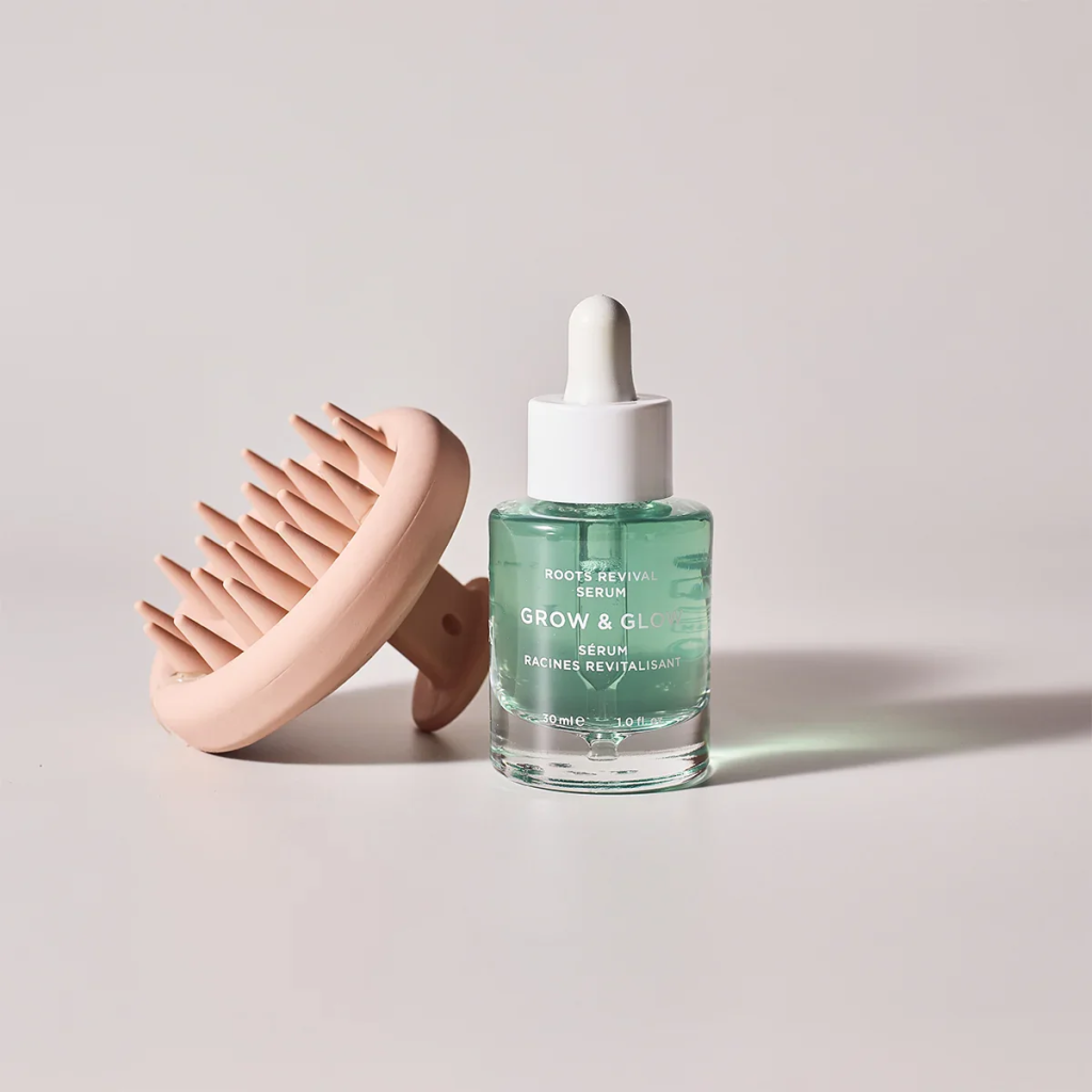 בקבוקון סרום GROW & GLOW לשורשי שיער עם פיפטה, מונח ליד מברשת עיסוי קרקפת בצבע ורוד.