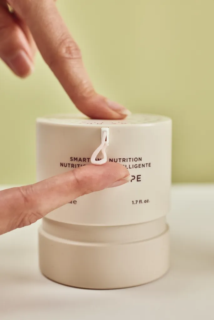 אישה מכניסה אצן עם העוזר במרחק לידה למיכל עם מעבדת טקסטיל. המיכל מצויד בהוראות שימוש וברקע קיר בצבע ירוק בהיר.