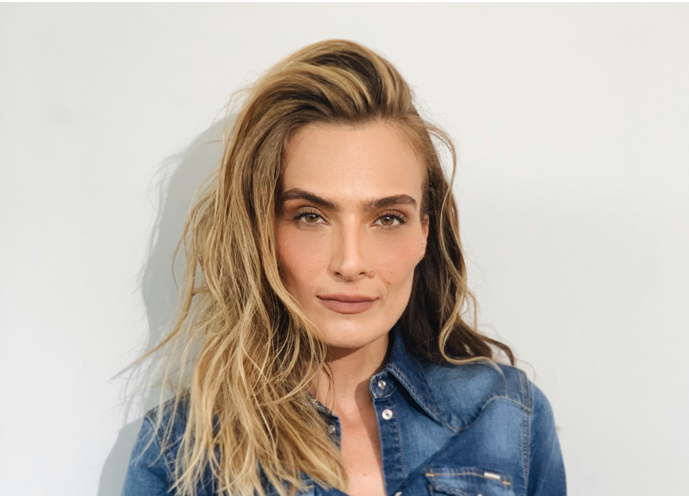 A woman with long, wavy hair wearing a denim jacket poses against a plain background.