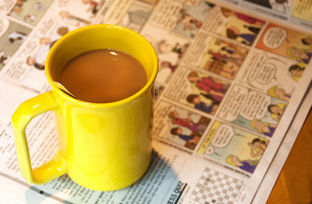 כוס צהובה מלאה קפה מונחת על עיתון עם קומיקס בצדדים.