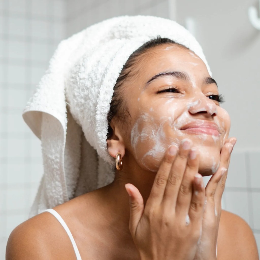 אישה שוטפת את פניה עם קצף ניקוי, לובשת טבעות ואחוזה במגבת על ראשה. היא מחייכת בזמן שהיא מנקה את עור פניה.