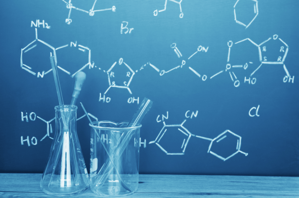 שני כלי זכוכית מעבדה מונחים על שולחן עץ, עם צינורות סיכון, על רקע קיר כחול עם נוסחאות כימיות ותרכובות כתובות בפתקיות לבנות.