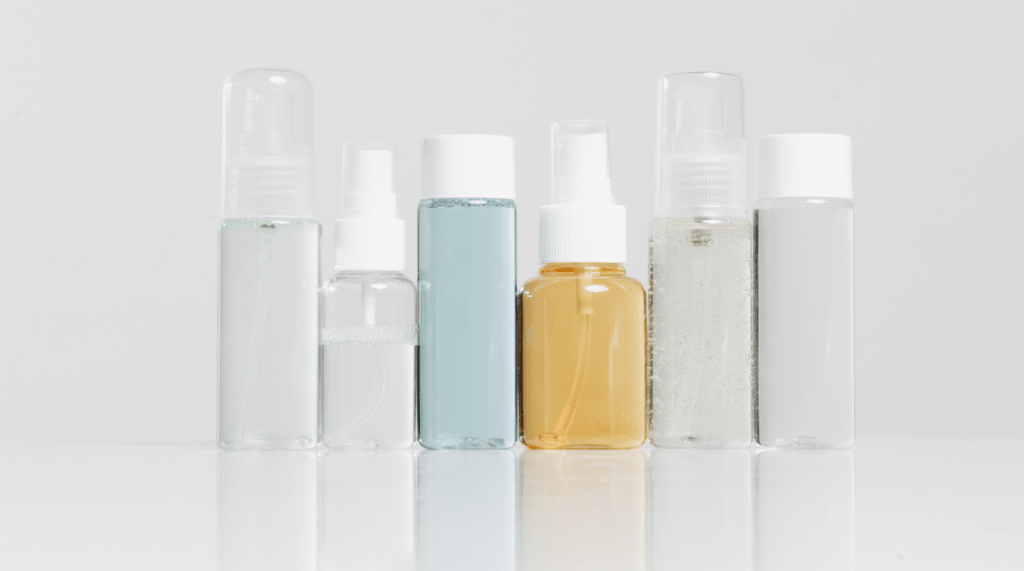 שישה בקבוקי תרסיסים ומיכלים שקופים שונים, מלאים בנוזלים שונים, עומדים על שולחן לבן עם רקע בהיר.