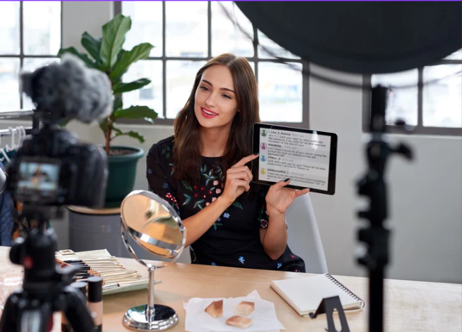 אישה יושבת עם טאבלט ומציגה תוכן לצלמת, עם מראה ומוצרי טיפוח על השולחן, ברקע צמח ירוק וחלונות גדולים.