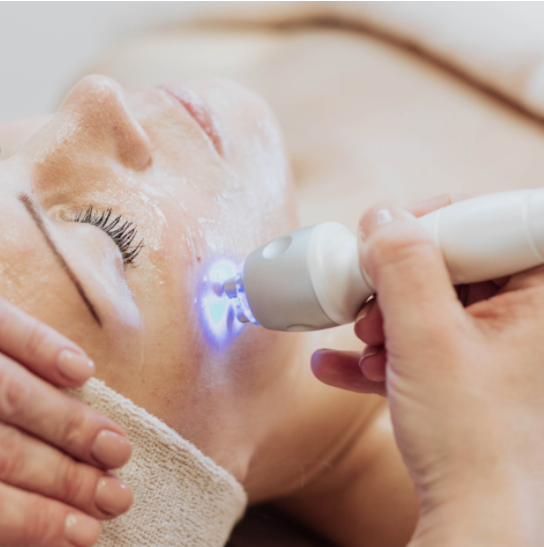 אישה שוכבת על מיטת טיפול עם חומרי טיפוח על פניה, בזמן שהמטפלת מבצעת טיפול בעזרת מכשיר emit אור כחול.